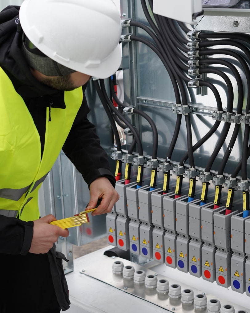 Ein:e Mitarbeiter:in steht mit Schutzhelm und gelber Warnweste vor einem offenen Schaltkasten eines Batteriegroßspeichers, in der Hand einen Zollstock.