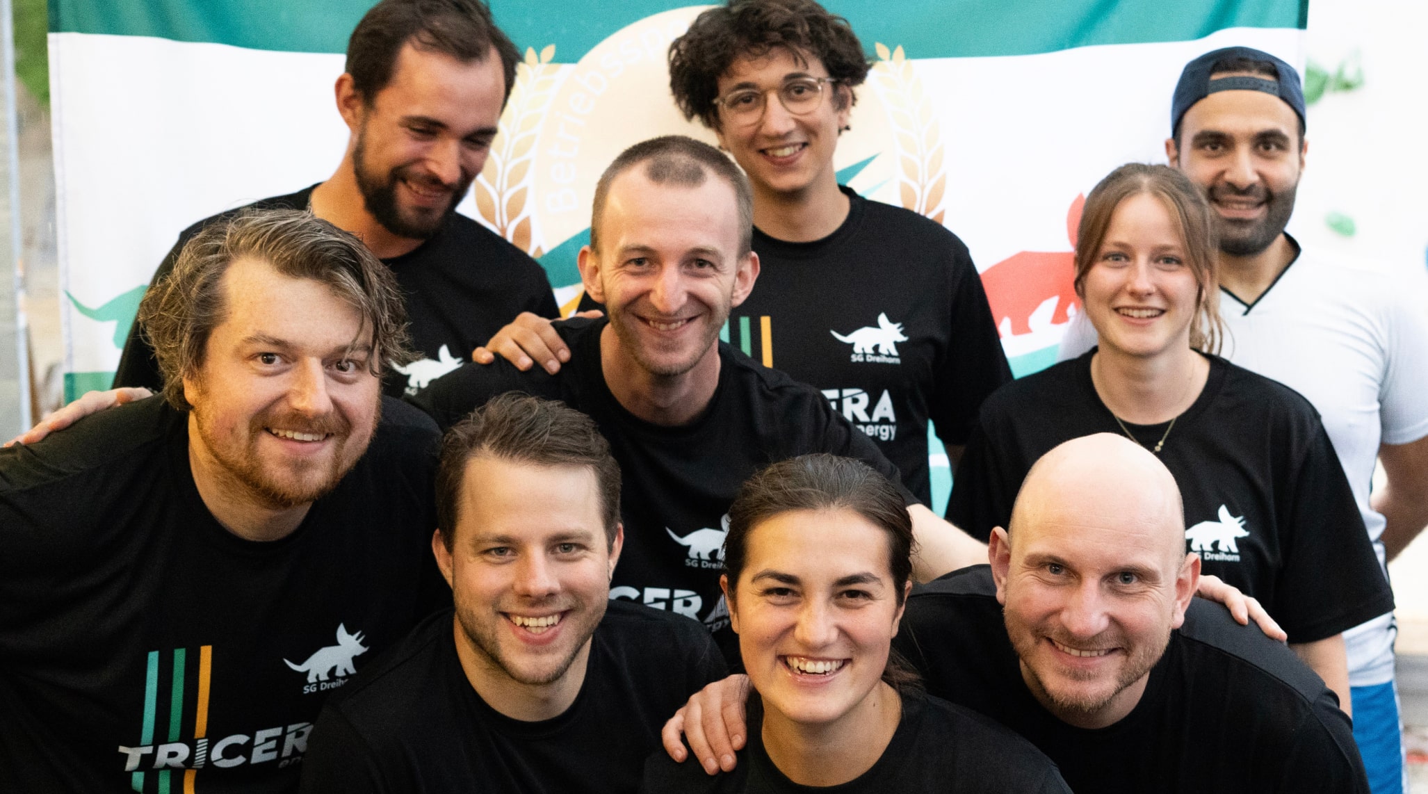 Neun Teammitglieder der Volleyball-Mannschaft des TRICERA SG Dreihorn lachen in die Kamera. Sie tragen die Team-T-Shirts mit einem aufgedruckten Triceratops.